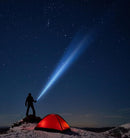 Person standing on a hilltop at night next to a lit tent shining a powerful MAGLITE flashlight beam into the starry sky
